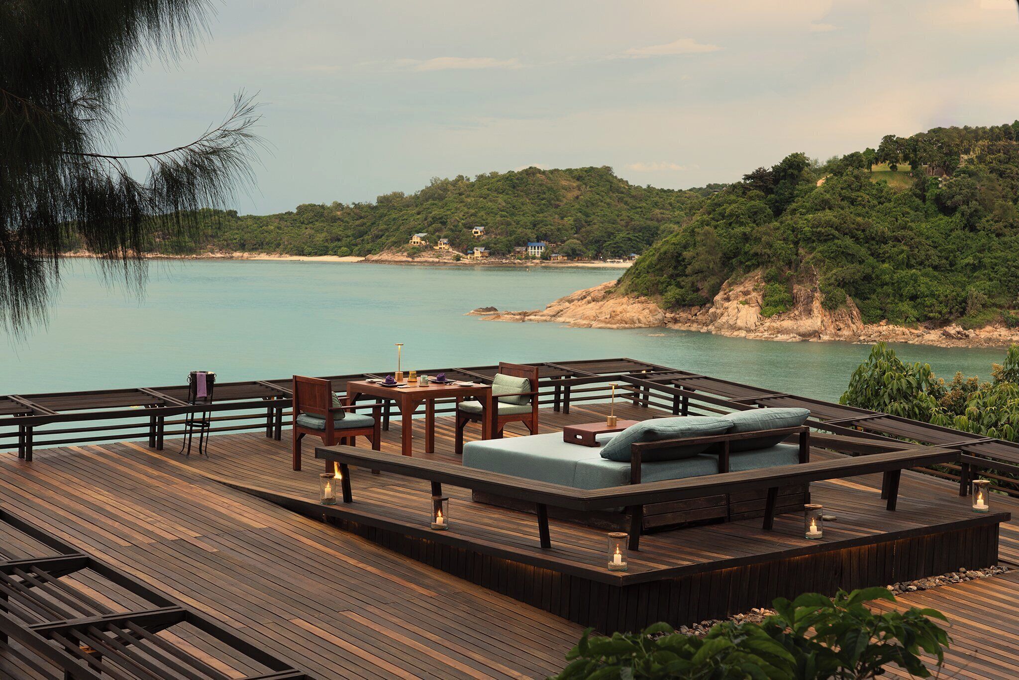 Una terraza de madera con mobiliario exterior, incluyendo un sofá y sillas, con vista a una masa de agua tranquila y una ladera boscosa.