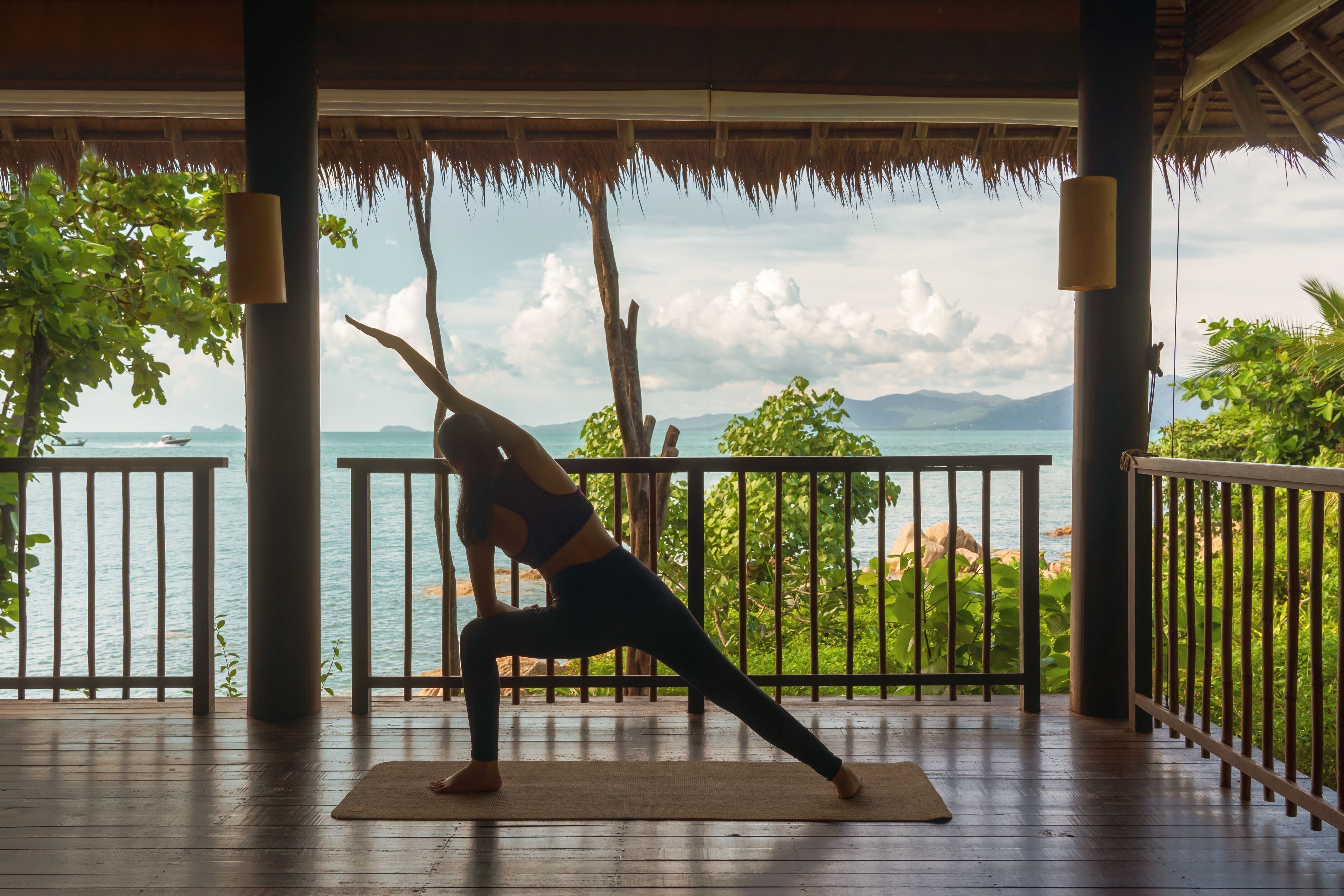 Una persona realizando una postura de yoga sobre una colchoneta en un espacio cubierto, con vistas al mar y a las montañas.