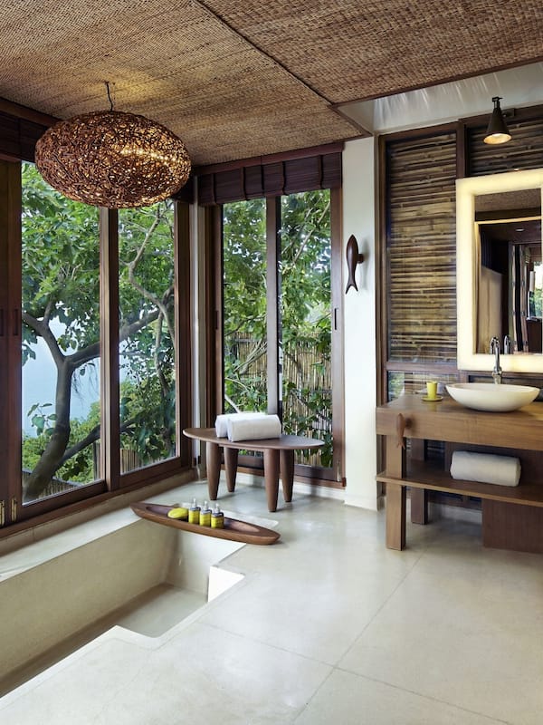 Sink, mirrors, and a woven pendant light adorn this bathroom, offering a view of lush greenery and a body of water.