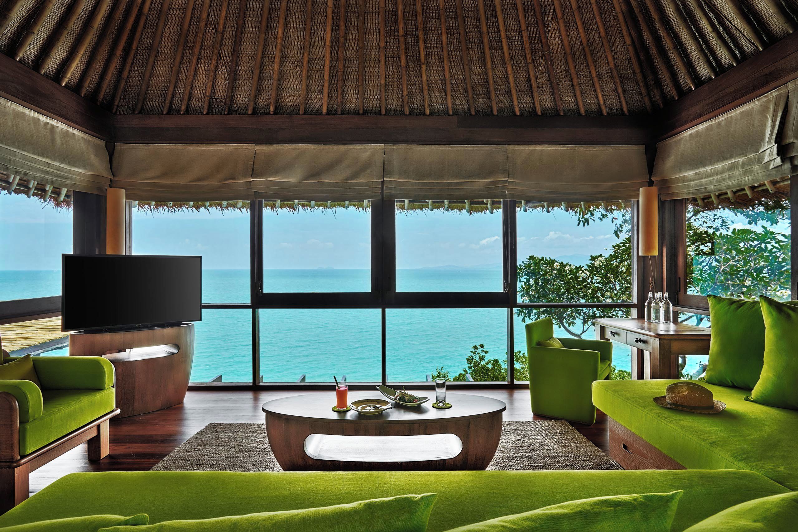 A room with a large window overlooking a body of water, a flat-screen TV, and green upholstered furniture.