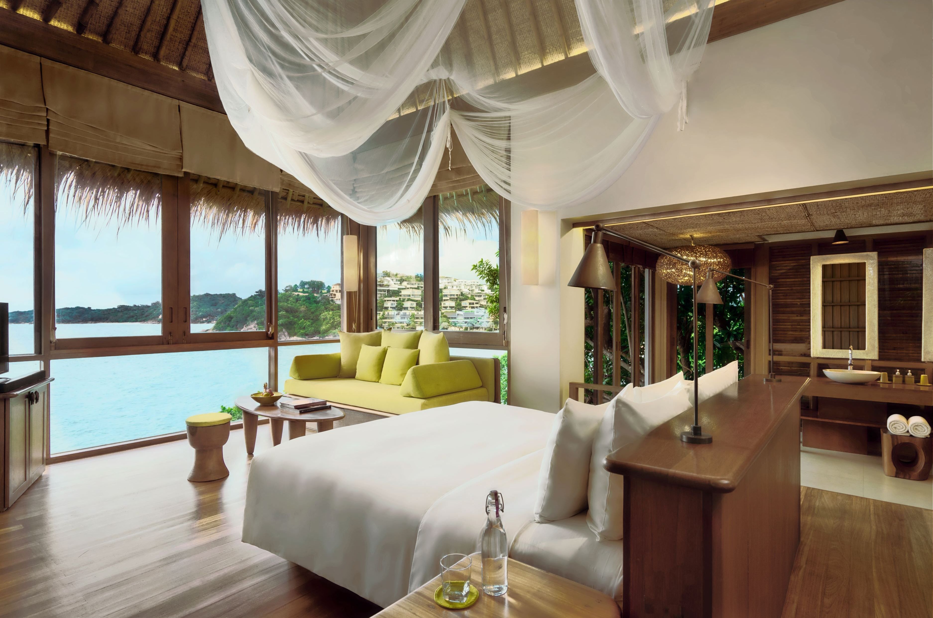 Une chambre spacieuse avec un grand lit, un coin salon avec un canapé jaune, un bureau en bois et une grande fenêtre donnant sur la mer.