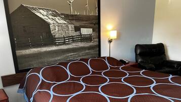 Een hotelkamer met een groot bed, een zwarte leren fauteuil, een wandlamp en een groot gepresenteerd schilderij van een schuur met windturbines.