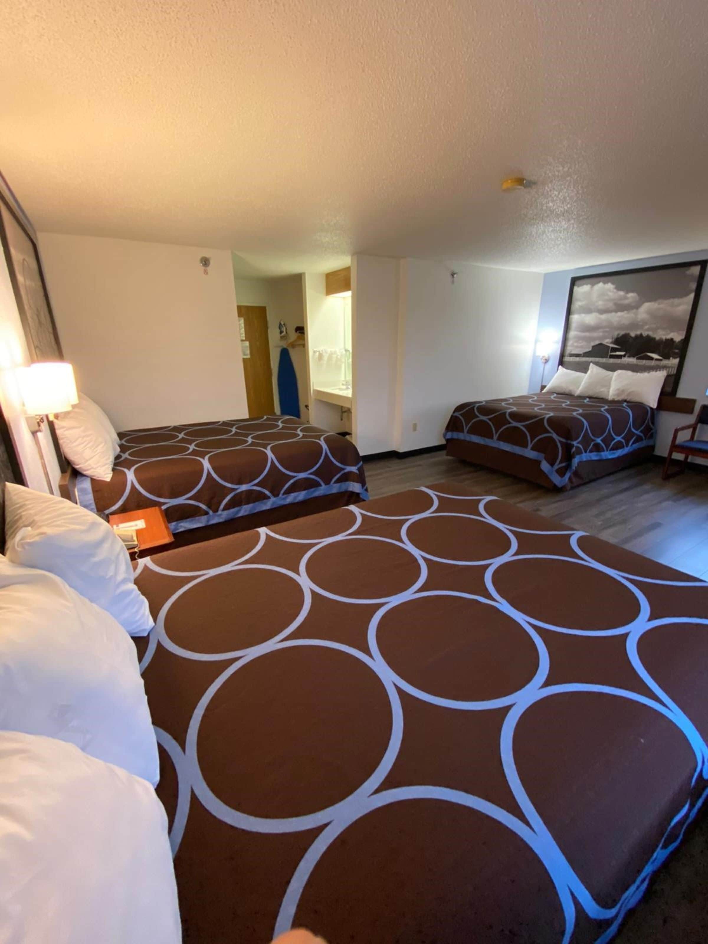 Habitación de hotel con tres camas, cada una con una colcha estampada. Se ve un ventanal grande a la derecha, una puerta que da a un baño y una mesita con una lámpara.