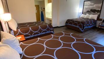 Habitación de hotel con tres camas, cada una con una colcha estampada. Se ve un ventanal grande a la derecha, una puerta que da a un baño y una mesita con una lámpara.
