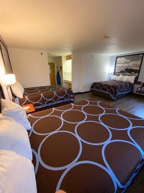 Habitación de hotel con tres camas, cada una con una colcha estampada. Se ve un ventanal grande a la derecha, una puerta que da a un baño y una mesita con una lámpara.