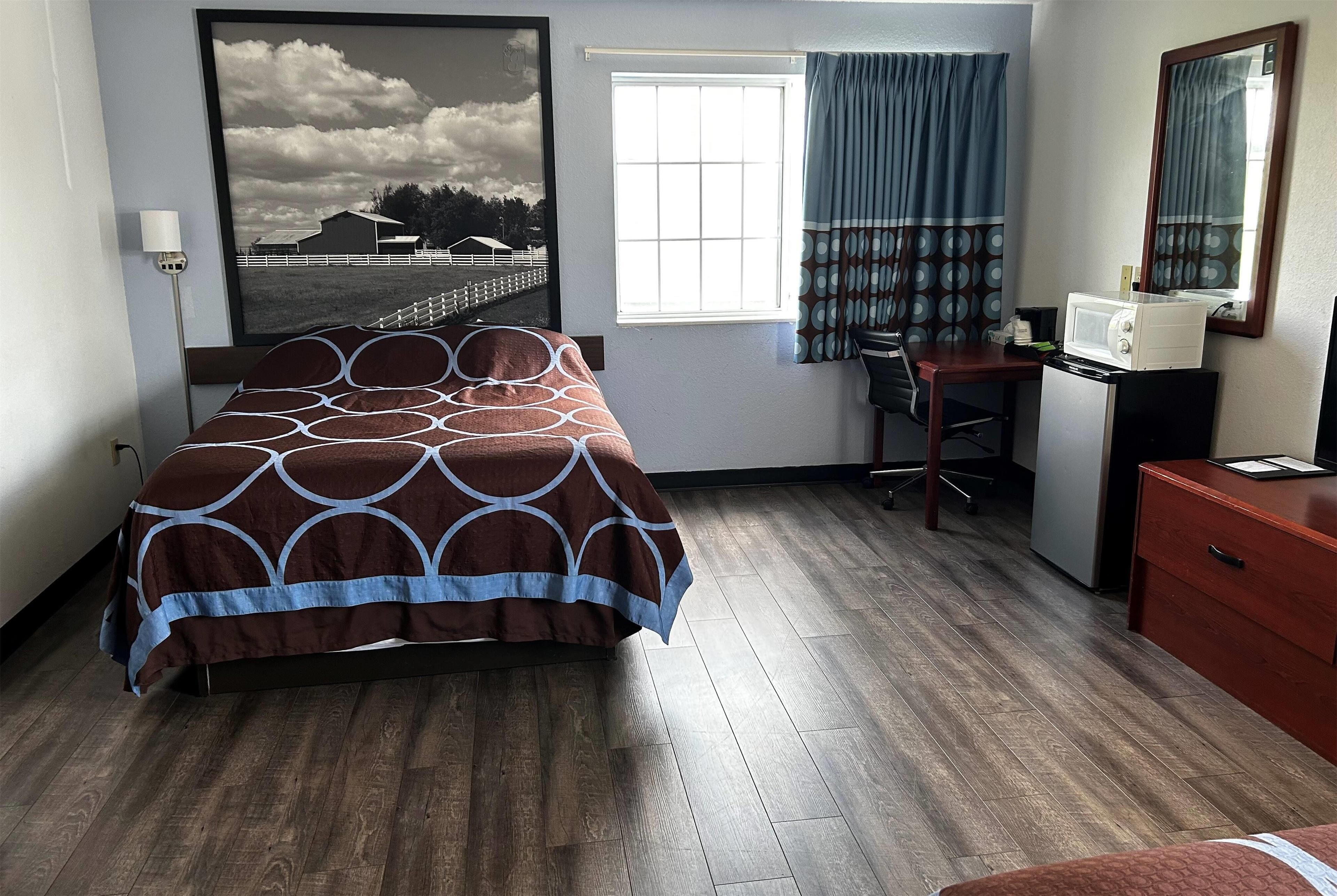 Habitación de hotel con cama, escritorio, silla, microondas y un amplio ventanal con cortinas.