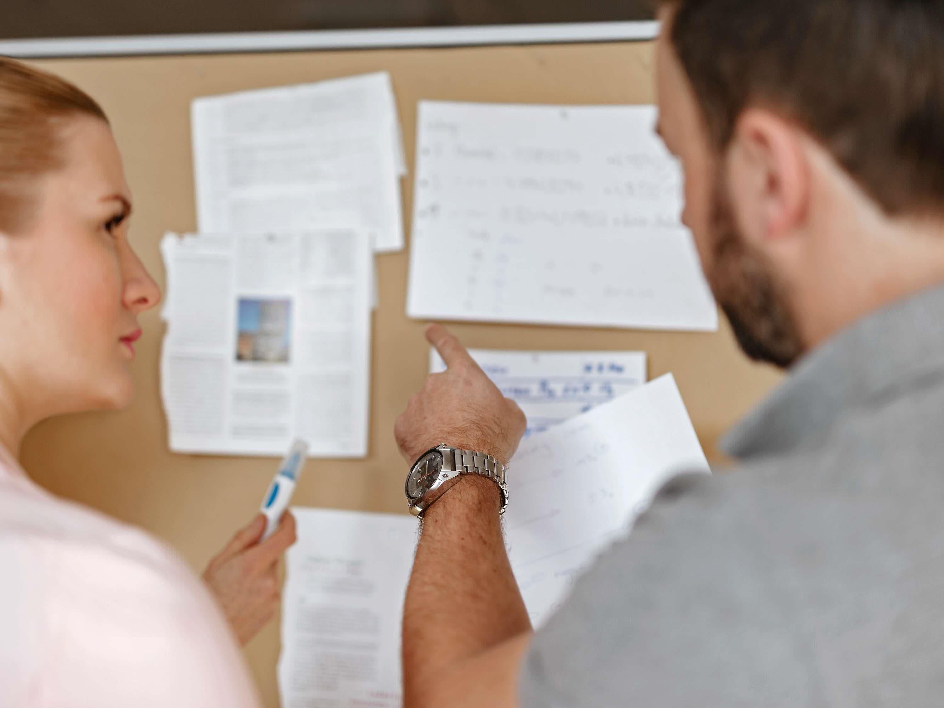 A person holding a pen and papers in front of a bulletin board with various documents pinned to it.
