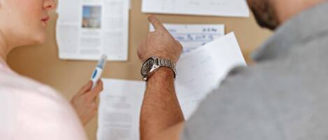 A person holding a pen and papers in front of a bulletin board with various documents pinned to it.