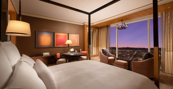 A hotel room with a large bed, a sitting area with a table and chairs, and a view of a cityscape at dusk. - Wynn Las Vegas (Las Vegas)