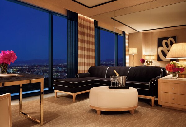 A modern living room with a large window, a black sofa, a white ottoman, a wooden side table, and a lamp.