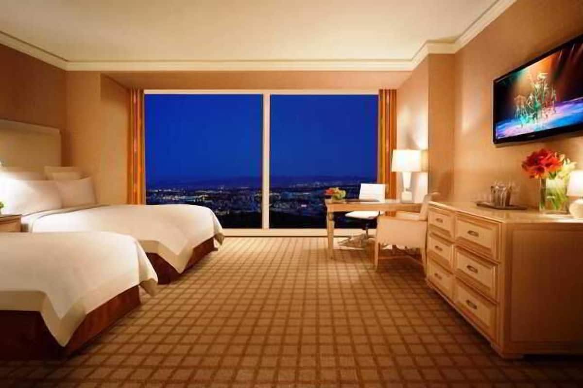 A hotel room with two beds, a desk, a TV, and a view of the city at night.