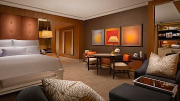 Habitación de hotel con una cama grande, un escritorio con una silla, un sofá y una mesita pequeña. Se observan cuadros enmarcados en la pared y una lámpara sobre el escritorio.