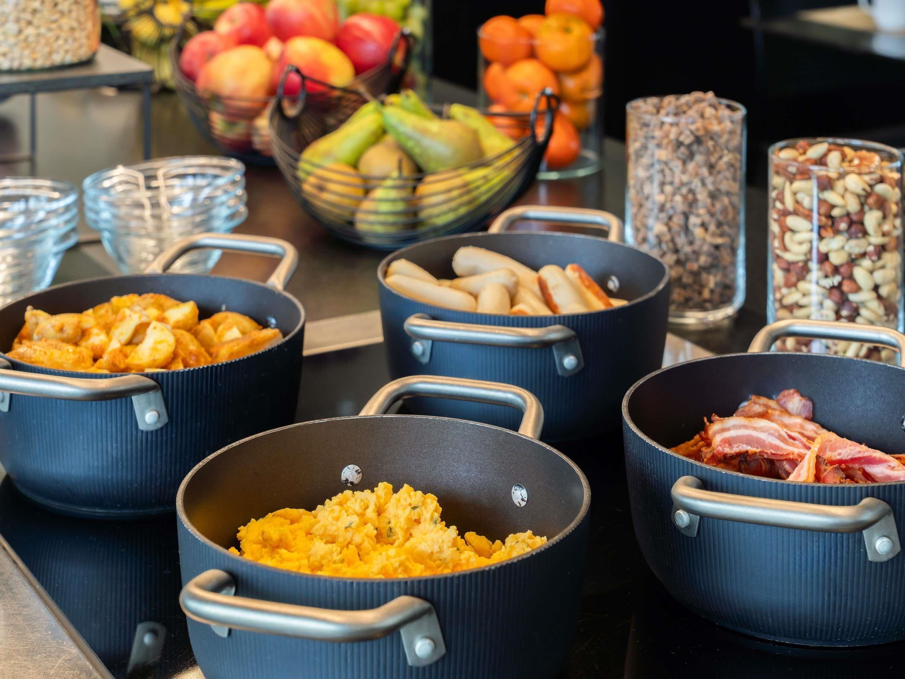 A kitchen counter with various cooking pots and pans, a bowl of fruits, and jars of nuts.