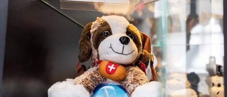 Un jouet pour chien en peluche orné d’un drapeau suisse, posé sur un objet bleu représentant un paysage urbain, derrière une vitrine.