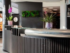 A reception desk with a modern design, featuring a glass top and a black, ribbed base. There are potted plants on the left side of the desk. - Novotel Suites Geneve Aeroport (Vernier)