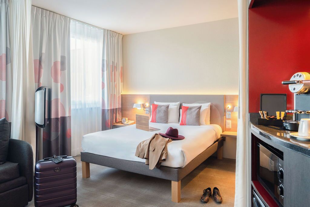 A hotel room with a bed, bedside tables, a desk, a chair, a suitcase, and a kitchenette.
