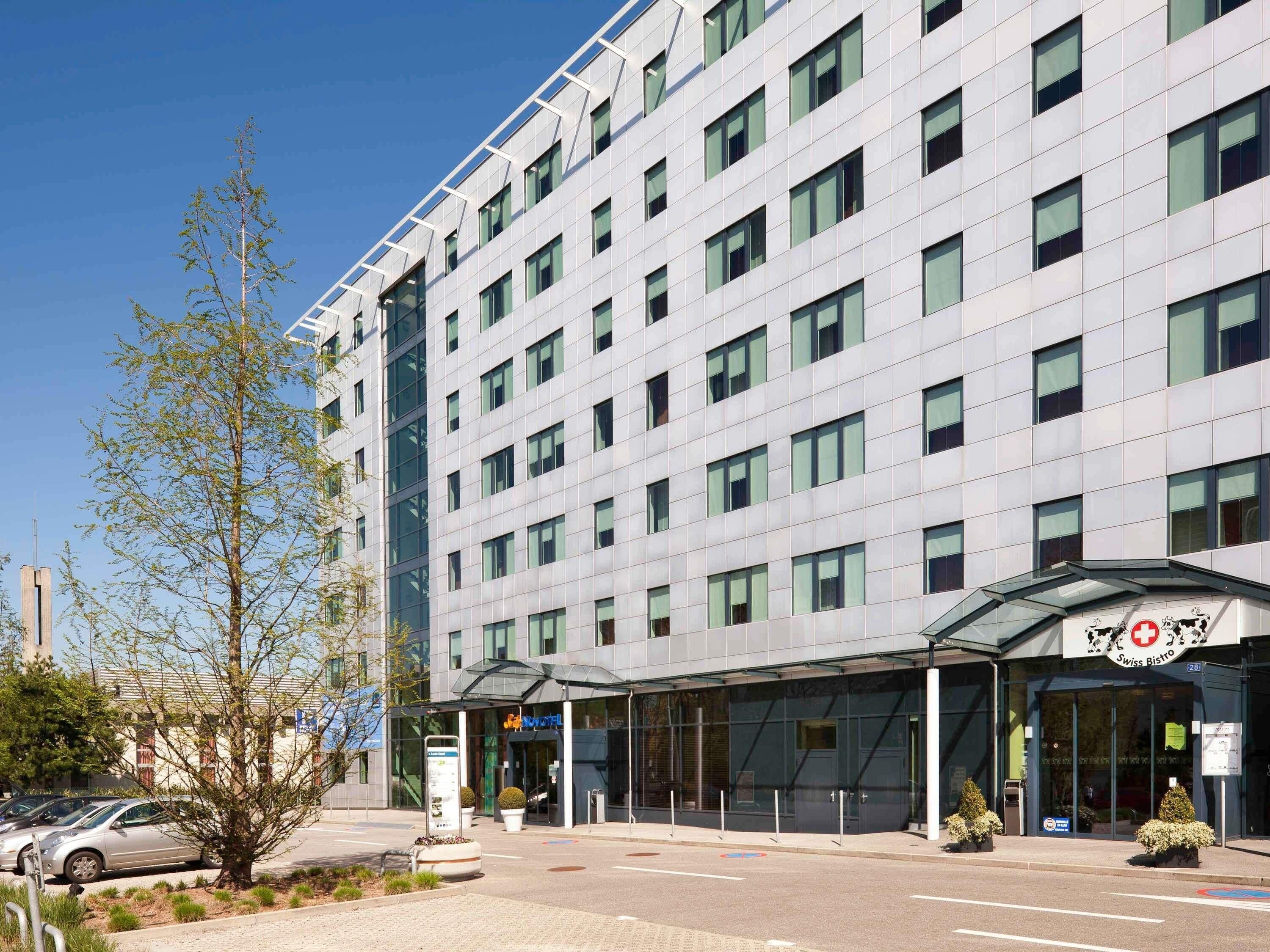 A modern hotel with a glass facade, a Swiss Bistro sign, and a parking area with cars and a tree.