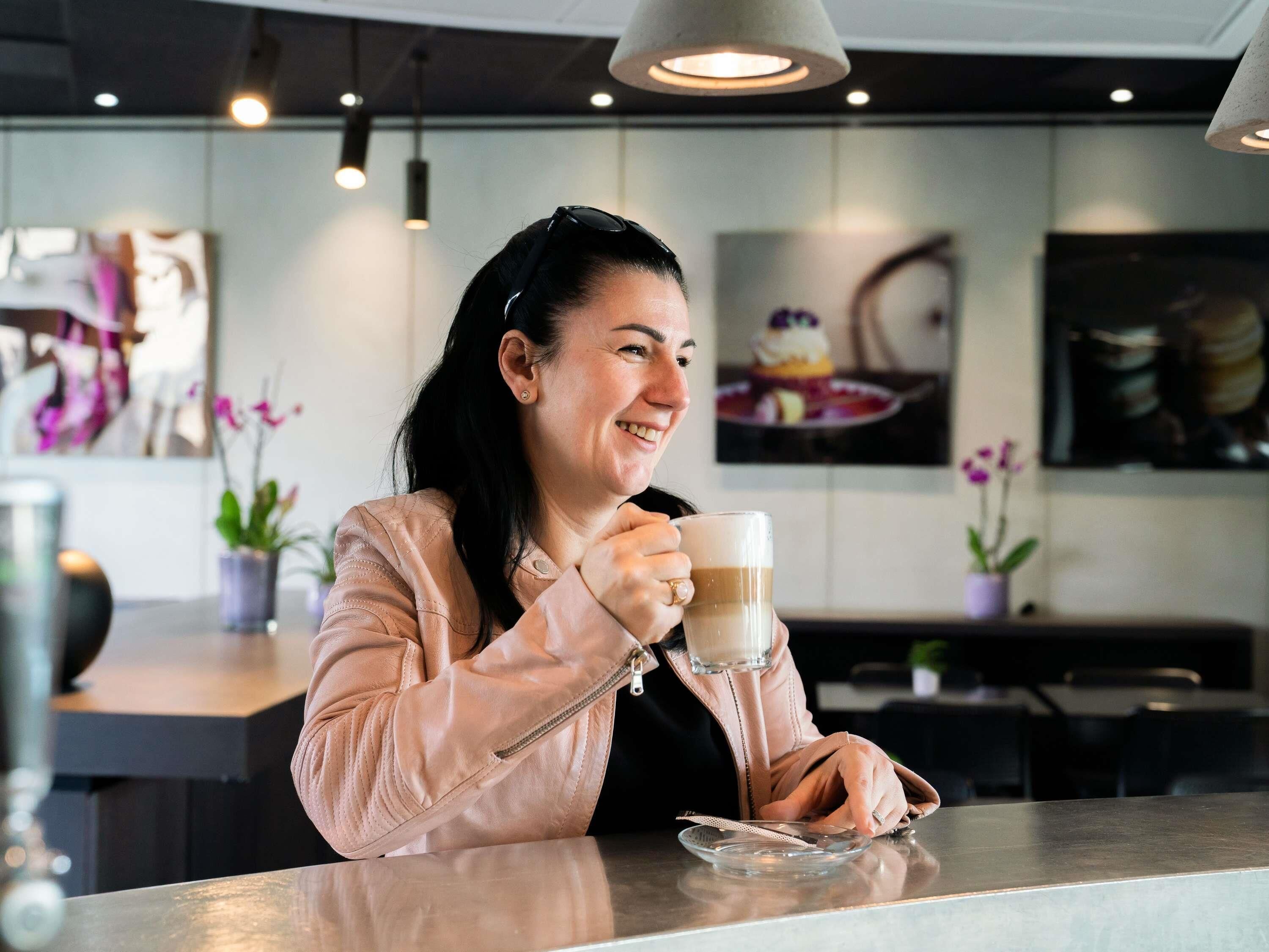 Une personne est assise à une table dans un café, avec une tasse de café devant elle. On aperçoit en arrière-plan un présentoir de desserts et des plantes en pot.
