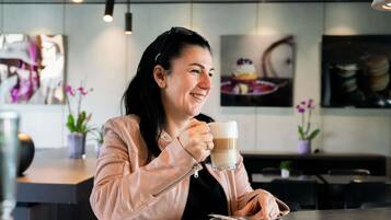 Une personne est assise à une table dans un café, avec une tasse de café devant elle. On aperçoit en arrière-plan un présentoir de desserts et des plantes en pot.