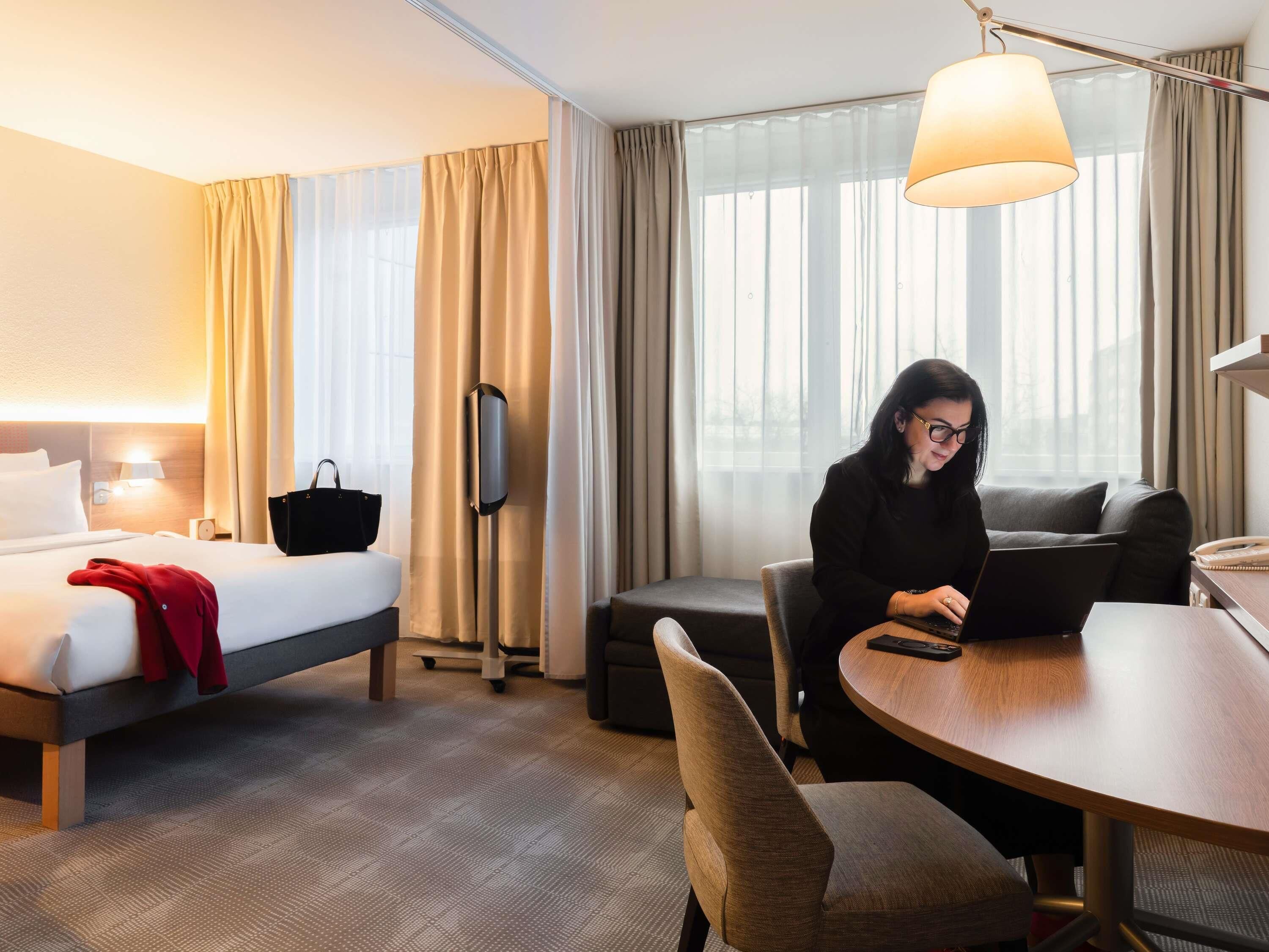 A hotel room with a bed, a desk, a chair, a lamp, and a TV.