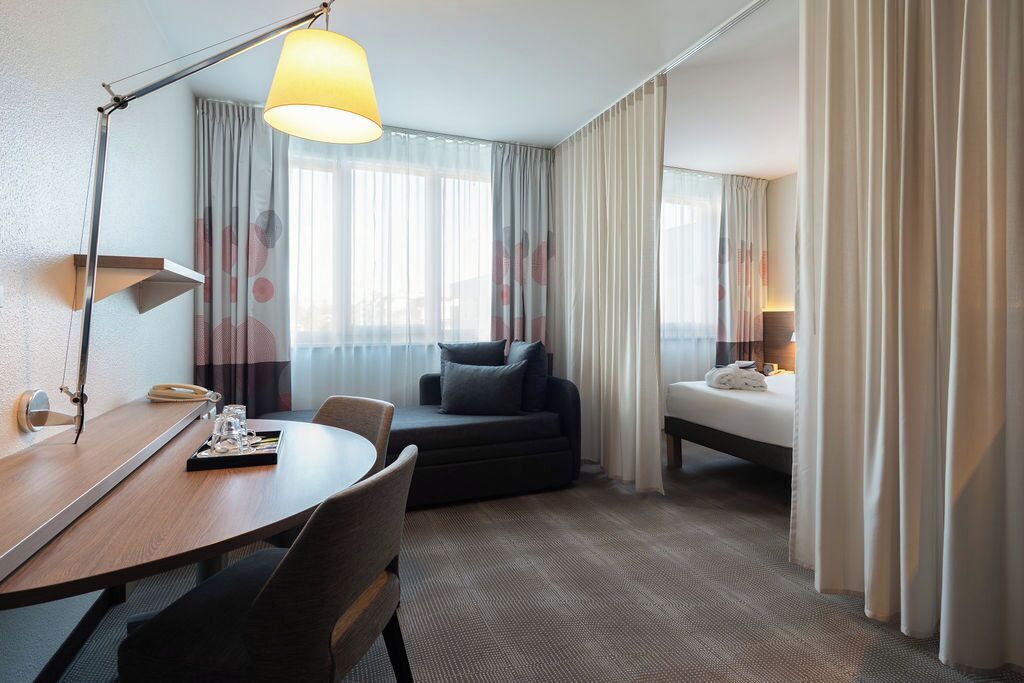 A hotel room with a round table, a sofa, a bed, and a desk with a lamp.