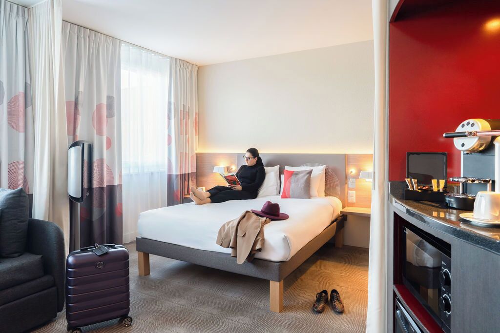 A hotel room with a bed, a desk, a chair, a suitcase, and a kitchenette.