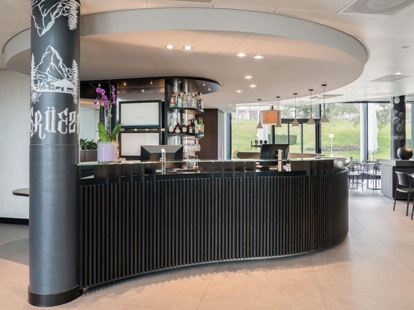A modern bar area with a curved counter, a large pillar with a mountain design, and a view of a green landscape through large windows. - Novotel Suites Geneve Aeroport (Vernier)