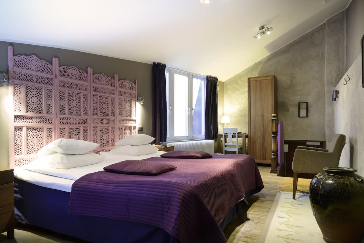 A hotel room with a large bed, a decorative headboard, a window with curtains, a wooden wardrobe, and a large vase on the floor.