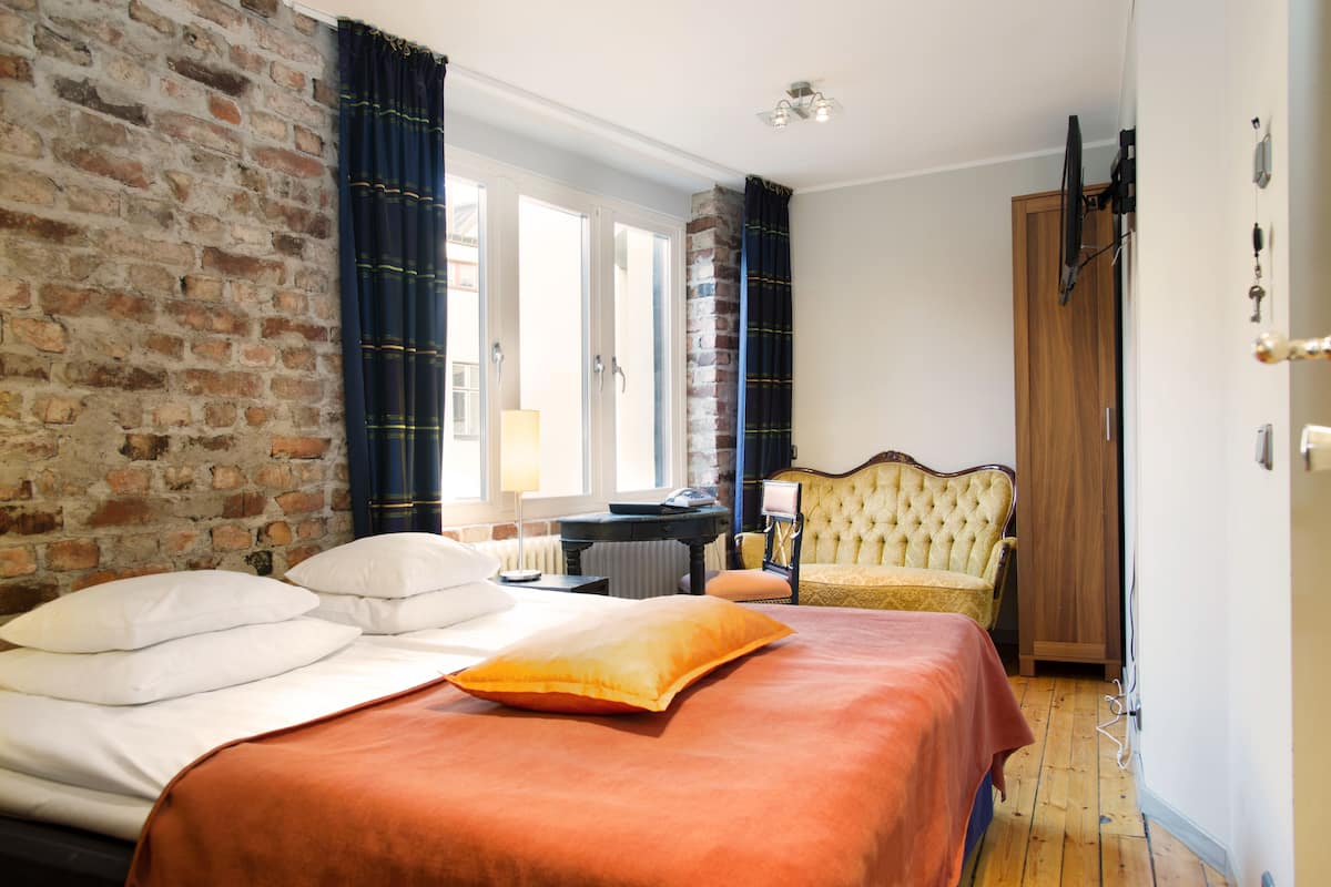 A hotel room with a brick wall, a bed with white and orange bedding, a wooden wardrobe, and a yellow armchair.
