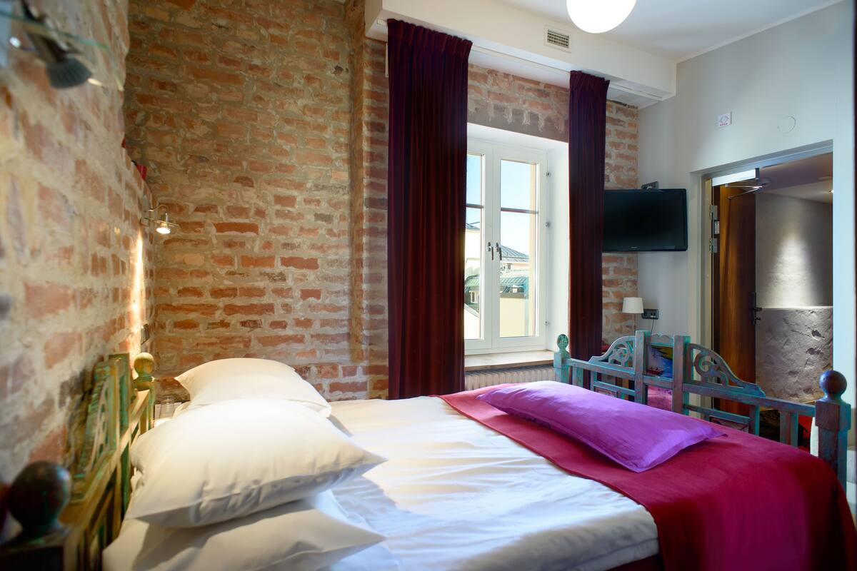 A room with a brick wall, a bed with white and purple bedding, a wooden headboard, and a TV mounted on the wall.