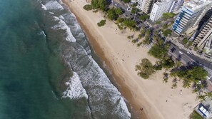 An aerial view of a beach with waves, palm trees, and buildings in the background.
