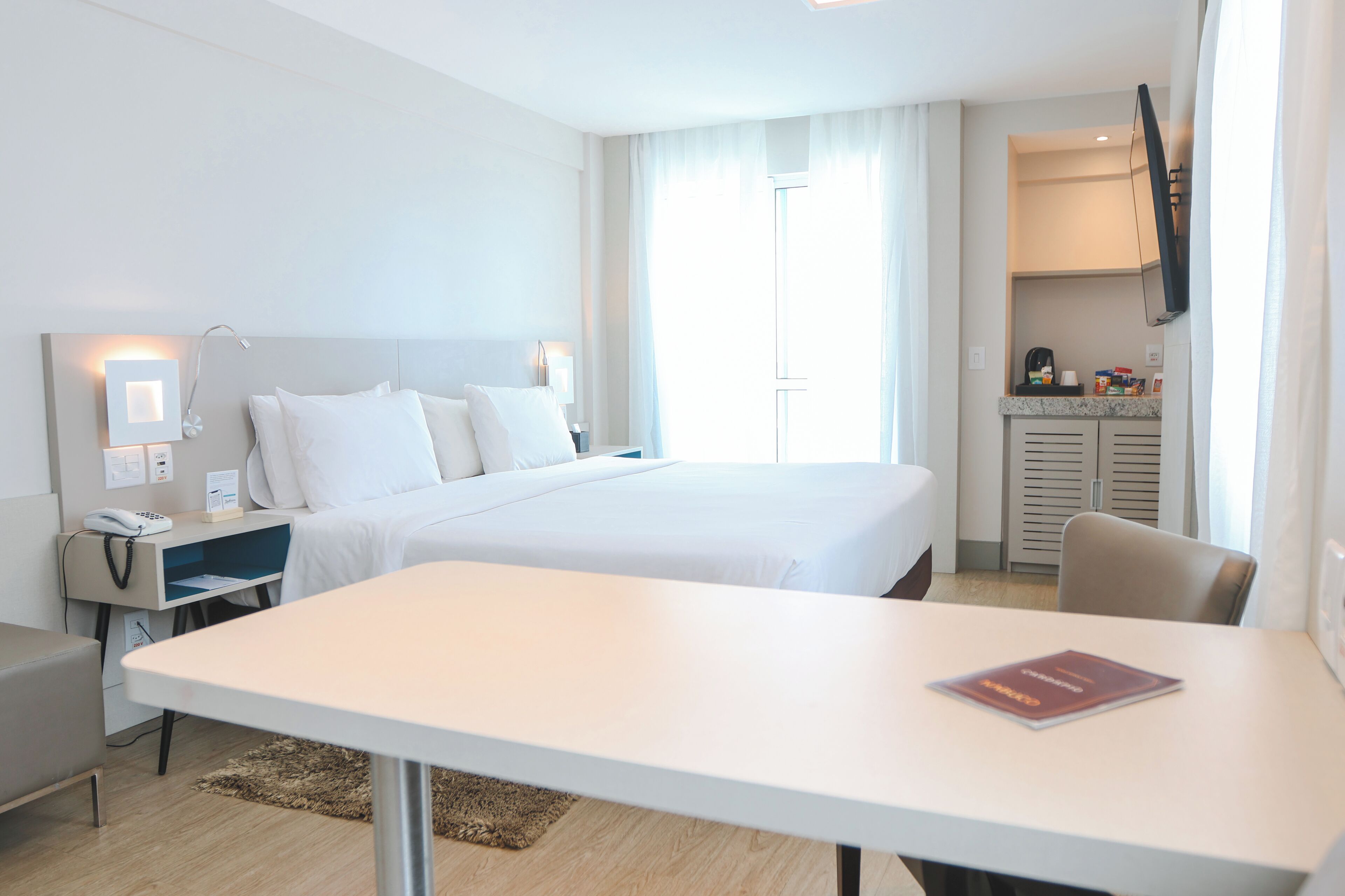 Une chambre d’hôtel comprenant un lit, des tables de chevet, un bureau, une chaise, un téléviseur fixé au mur et une fenêtre avec des rideaux.