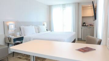 Quarto de hotel com cama, mesas de cabeceira, escrivaninha, cadeira, televisão na parede e janela com cortinas.