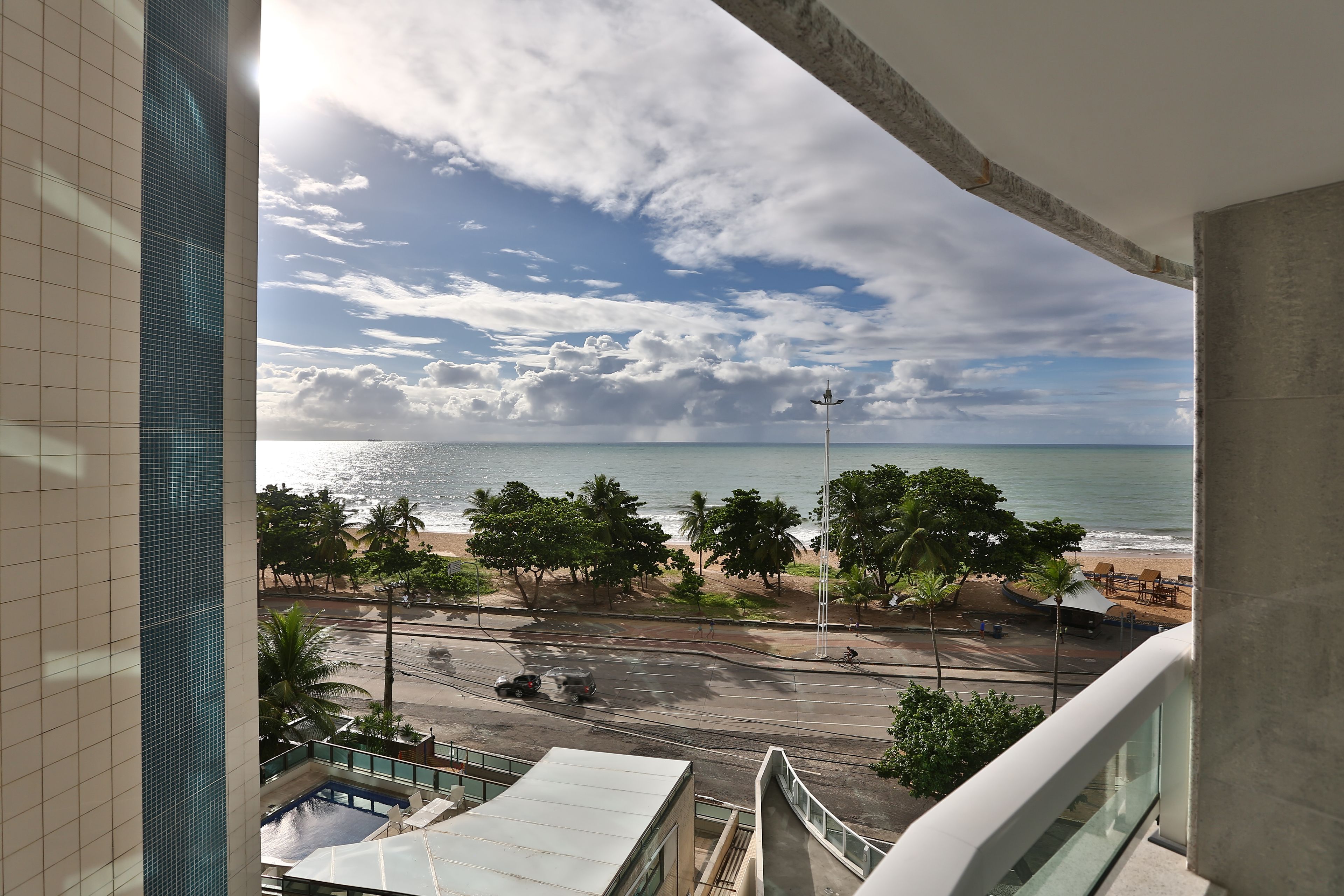 A view from a balcony overlooking a beach with palm trees, a road, and a building with a tiled facade.