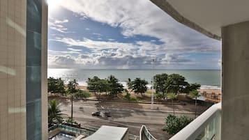 A view from a balcony overlooking a beach with palm trees, a road, and a building with a tiled facade.