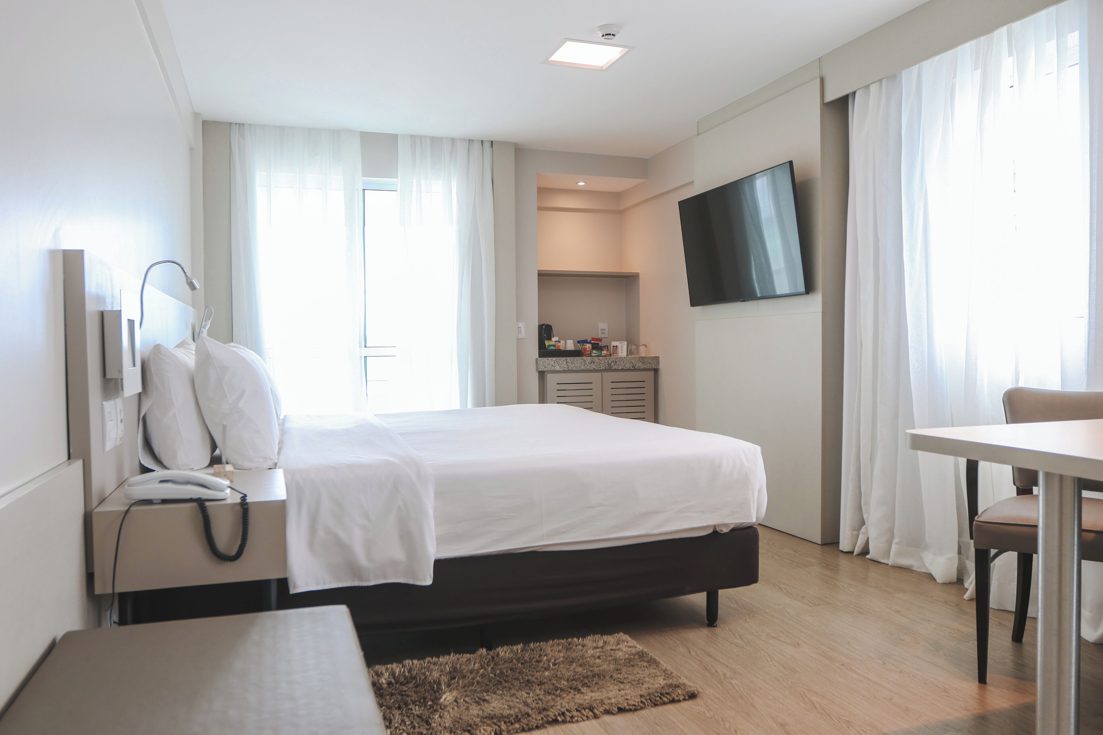 A hotel room with a bed, bedside table, lamp, phone, TV, and a small kitchenette area.