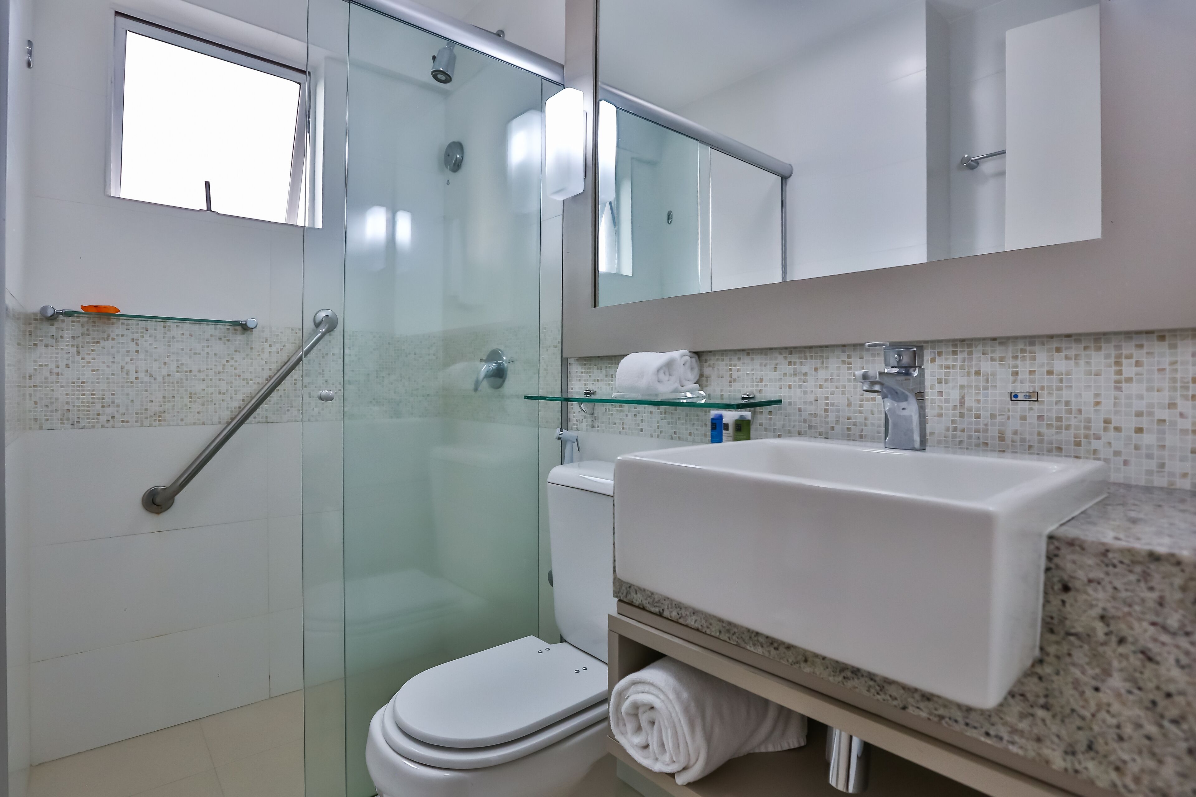 Un baño moderno con cabina de ducha de vidrio, inodoro blanco, lavabo con estante de vidrio y espejo sobre el lavabo.