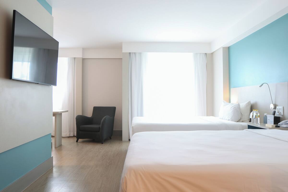 Ein Hotelzimmer mit zwei Betten, einem an der Wand montierten Fernseher, einem Sessel, einem Nachttisch mit Lampe und einem Fenster mit Vorhängen.