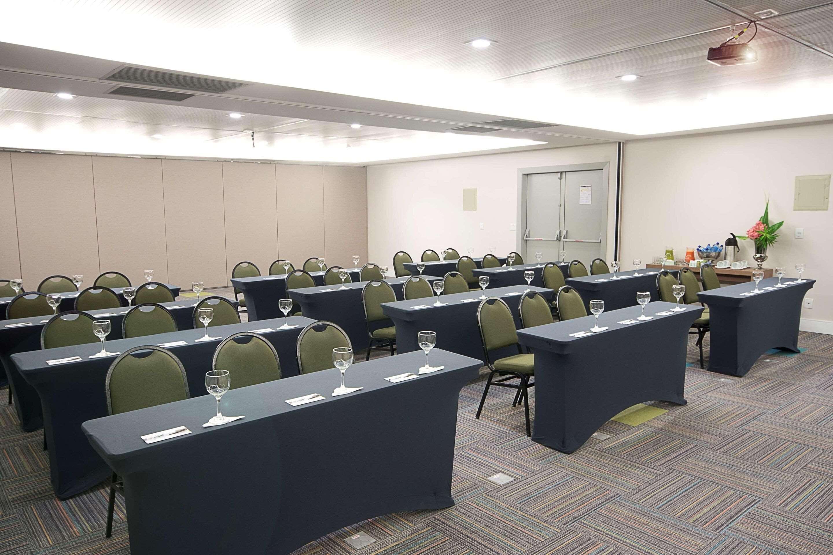Sala de conferências com várias mesas, cada uma com um copo de água e um bloco de notas. O piso é coberto por carpete e há um projetor no teto.