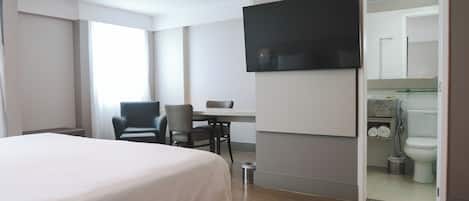Habitación de hotel con cama, escritorio, silla, televisión montada en la pared y baño visible a través de una puerta abierta.