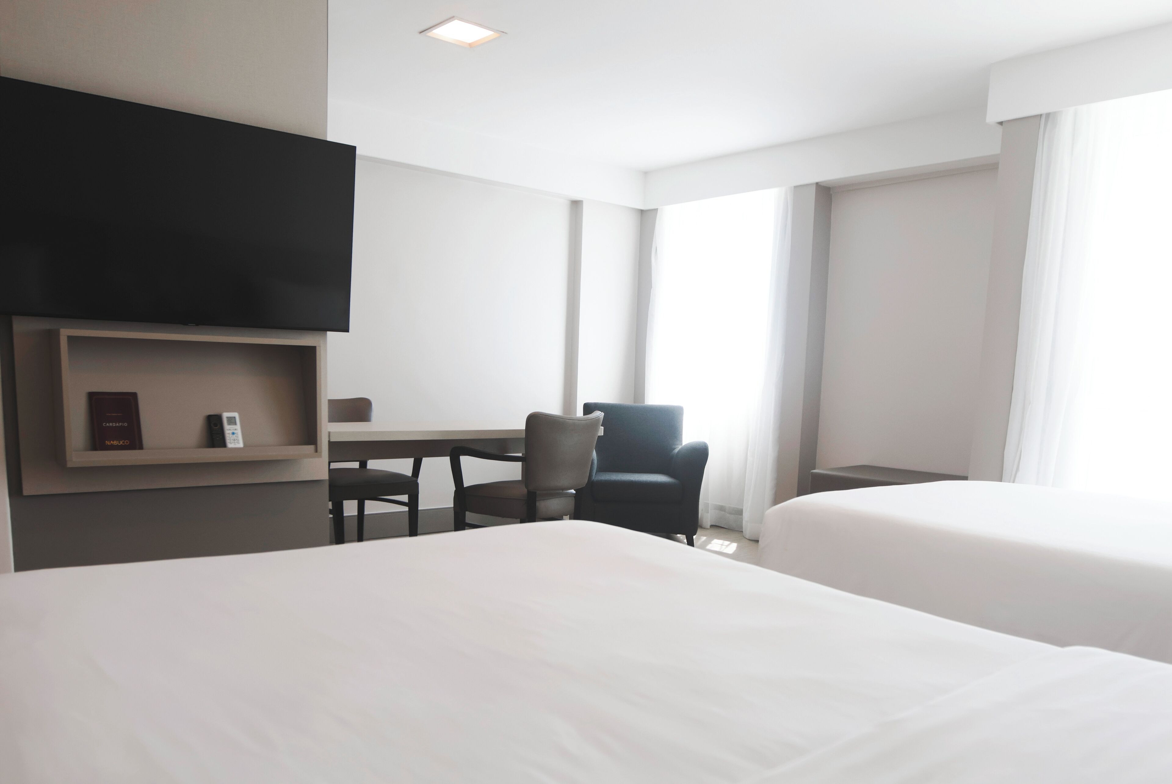 A hotel room with two beds, a desk, a chair, a TV mounted on the wall, and large windows.