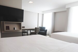 Habitación de hotel con dos camas, un escritorio, una silla, televisor montado en la pared y ventanas amplias.