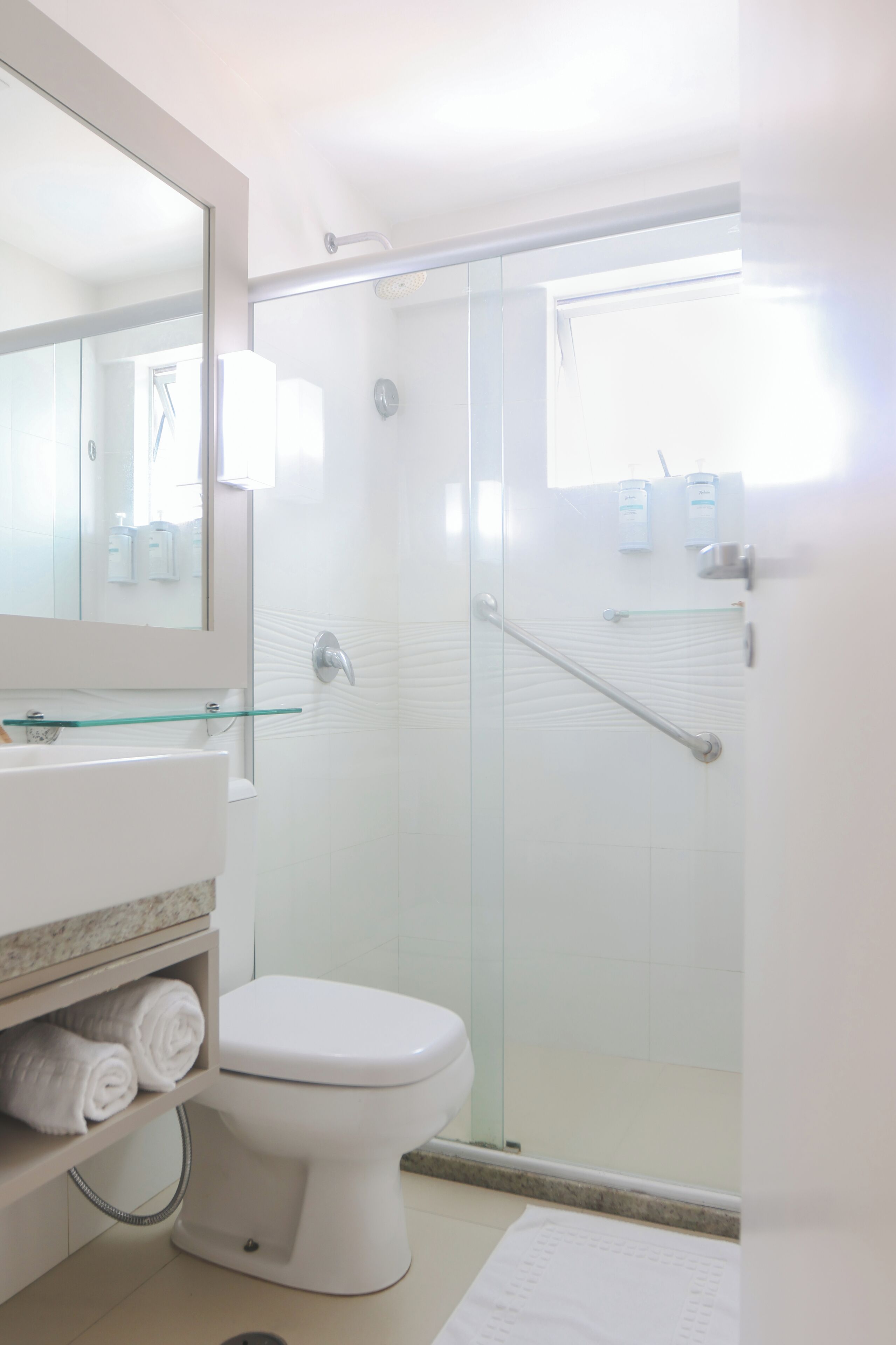Een moderne badkamer met een douchecabine van glas, een wit toilet, een glazen plankje met toiletartikelen en een grote spiegel.