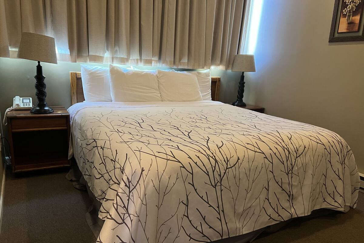 Uma cama bem arrumada com colcha estampada com padrão de galhos de árvore, duas luminárias de cabeceira e um telefone ao lado da cama em uma mesa de madeira.