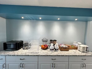 A kitchen with a microwave, toaster, clear storage box, fruit bowl, coffee makers, and a bread basket.