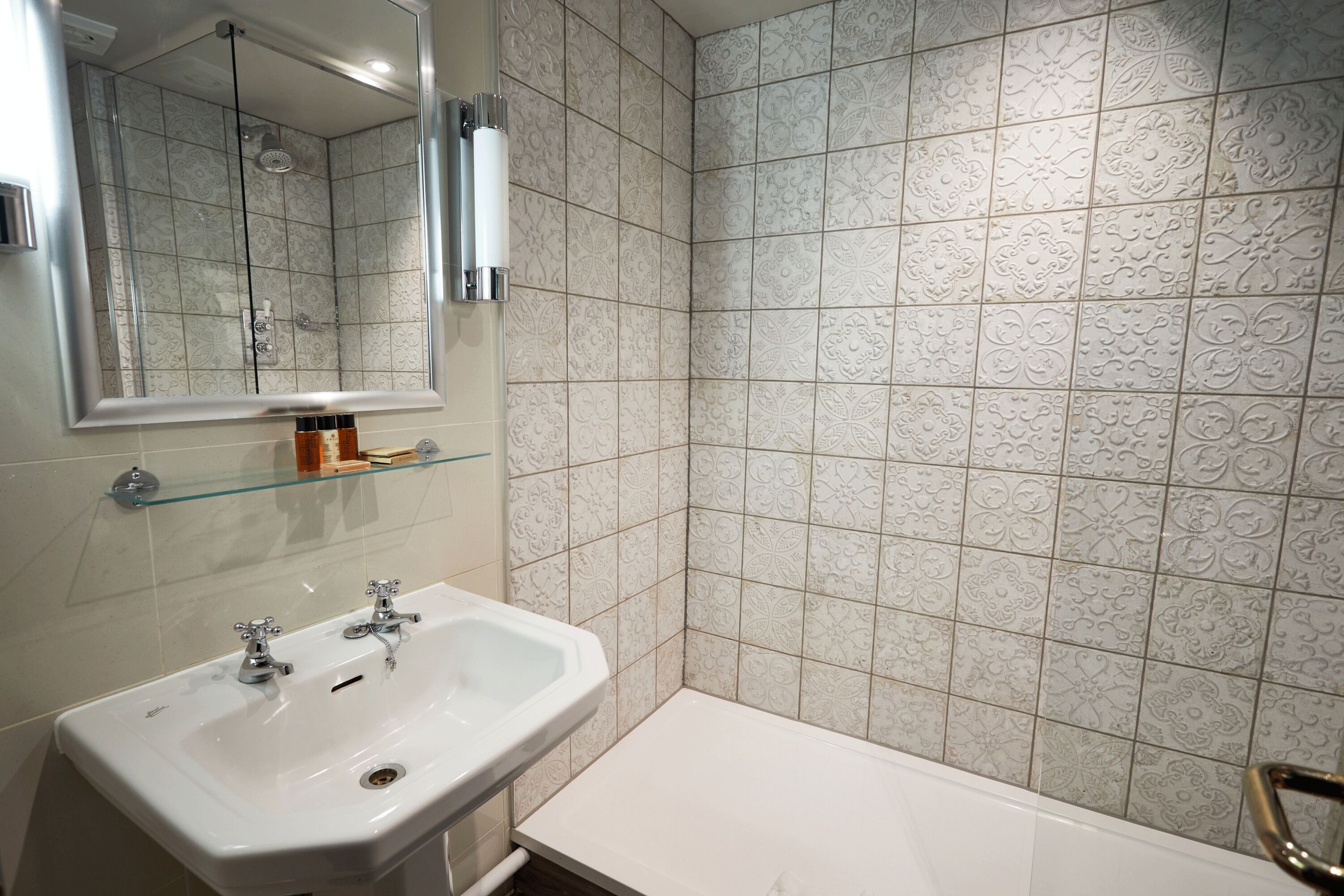 A bathroom with a white sink, a glass shelf, a mirror, and patterned tiles.