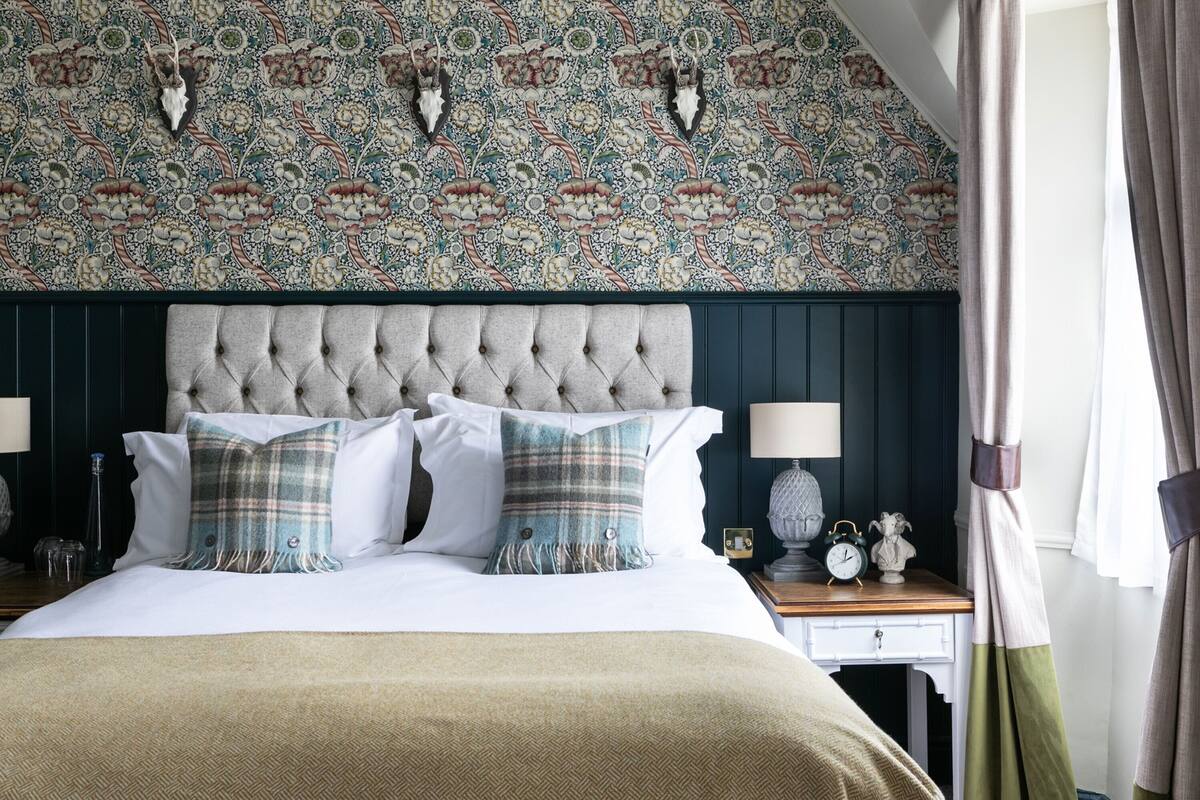 A bedroom with a tufted headboard, a patterned wallpaper, and a bedside table with a clock and a lamp.