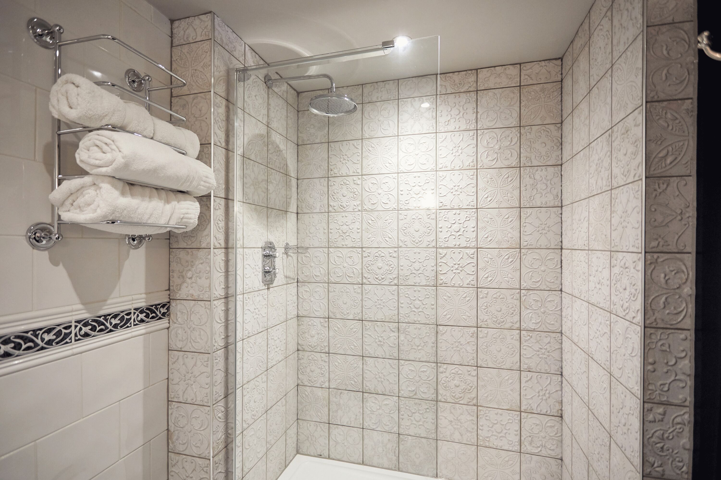 A shower area with a glass door, a towel rack with folded towels, and patterned tiles on the walls.