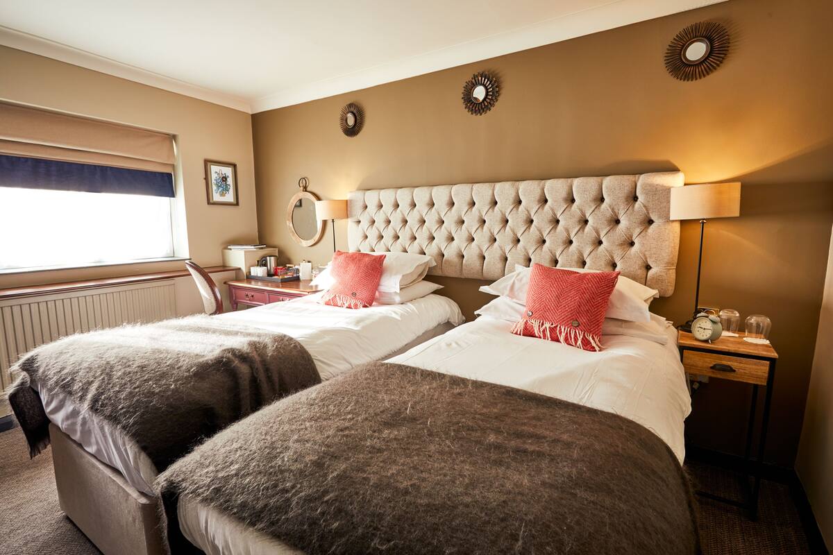 A hotel room with two beds, a large tufted headboard, bedside lamps, a nightstand, and a window with a view.