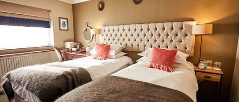 A hotel room with two beds, a large tufted headboard, bedside lamps, a nightstand, and a window with a view.